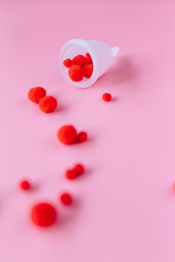 White silicone menstral cup on a plain pink background containing small red pom poms spilled ad trailing down the image.