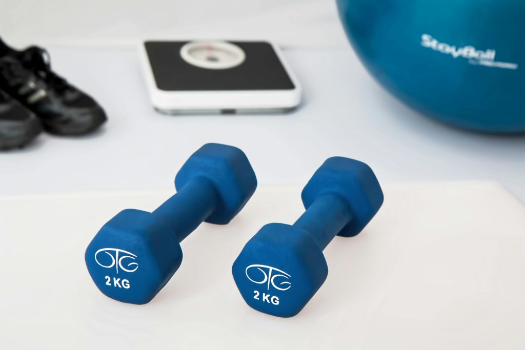 A pair of blue 2kg handheld dumbbell on a white floor in front of a pair of weighing scales, black workout shoes and a blue exercise ball