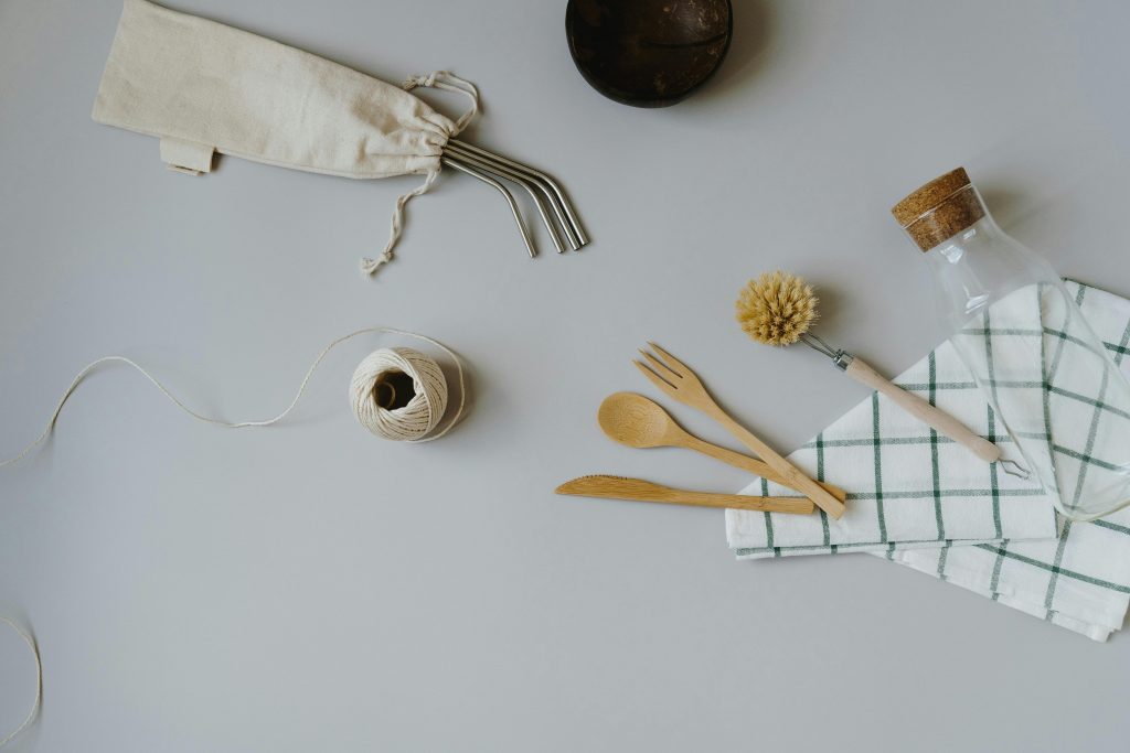 Flat lay image of the following items - clockwise from the top. a small dark copper dish,, a glass drinking bottle topped with a large cork stopper, next to a bamboo bottle cleaning brush, wooden cutlery, a ball of natural twine, a pack of reusable metal straws in a small linen bag