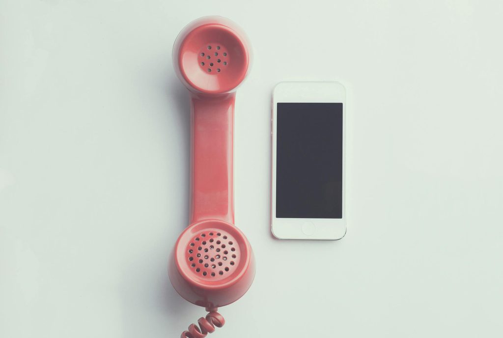 Red corded telephone handset next to a white iPhone on a plain white background