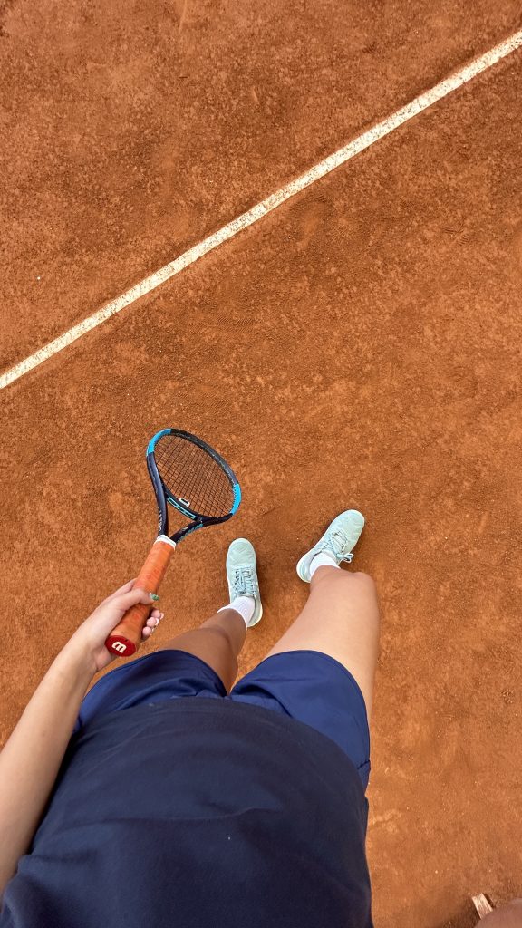Overhead picture of a woman taken from the torso down. She is standing on an orange tennis court, wearing a pair of navy blue shorts and light blue tennis shoes and holding a tennis racquet in her left hand.